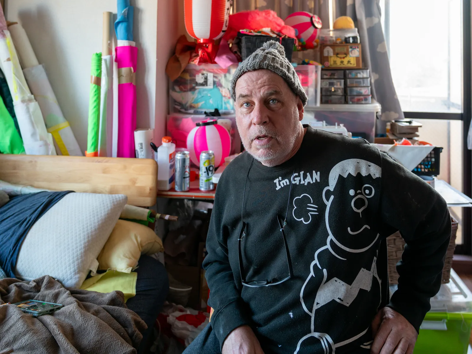 An expressive environmental portrait capturing the artist within his dense workspace, surrounded by vibrant neon textiles and traditional Japanese artifacts. The composition highlights cross-cultural themes and the tactile nature of his creative practice, creating a mood of creative intensity and cultural synthesis.