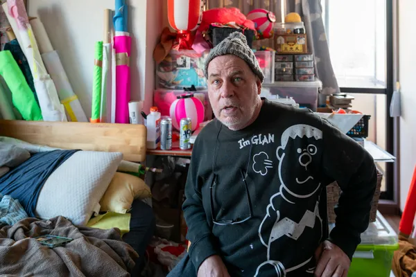 An expressive environmental portrait capturing the artist within his dense workspace, surrounded by vibrant neon textiles and traditional Japanese artifacts. The composition highlights cross-cultural themes and the tactile nature of his creative practice, creating a mood of creative intensity and cultural synthesis.