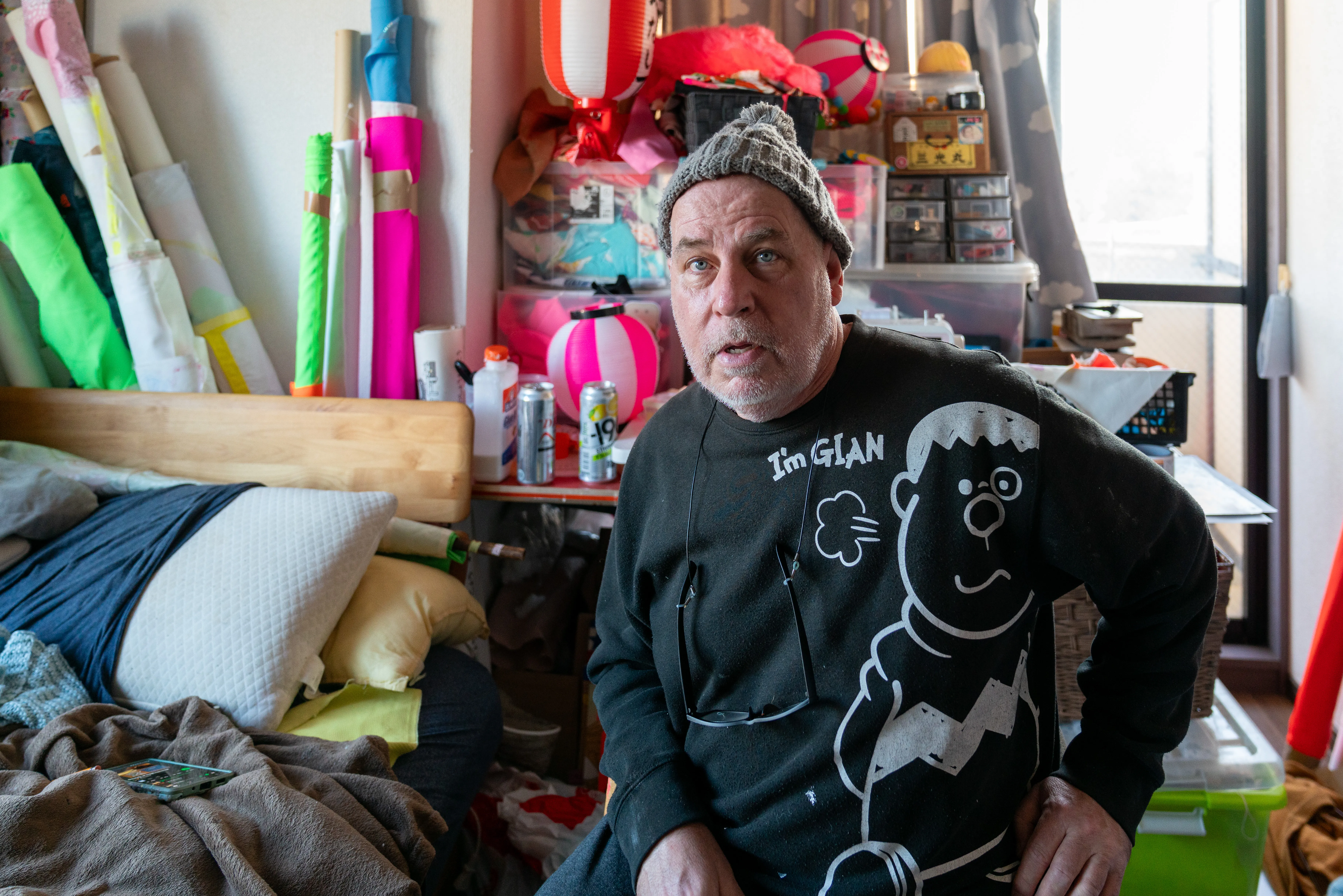 An expressive environmental portrait capturing the artist within his dense workspace, surrounded by vibrant neon textiles and traditional Japanese artifacts. The composition highlights cross-cultural themes and the tactile nature of his creative practice, creating a mood of creative intensity and cultural synthesis.