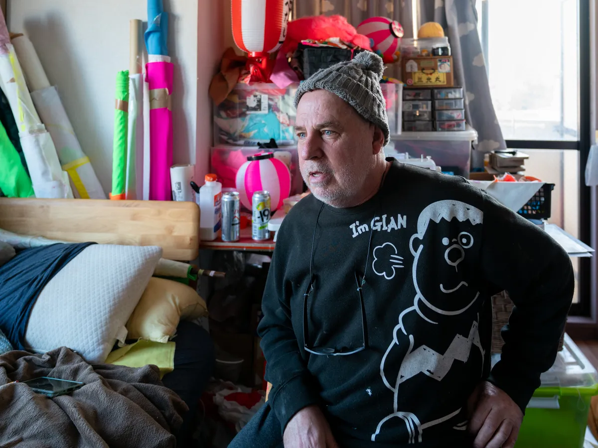 An environmental portrait of artist Dave Sayre in his cluttered workspace, surrounded by vibrant fabrics, Japanese lanterns, and various art supplies. The photograph captures a sense of creative immersion and the cross-cultural influences inherent in his life and work in Japan.