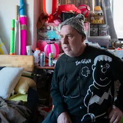 An environmental portrait of artist Dave Sayre in his cluttered workspace, surrounded by vibrant fabrics, Japanese lanterns, and various art supplies. The photograph captures a sense of creative immersion and the cross-cultural influences inherent in his life and work in Japan.