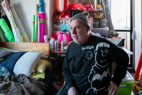 An environmental portrait of artist Dave Sayre in his cluttered workspace, surrounded by vibrant fabrics, Japanese lanterns, and various art supplies. The photograph captures a sense of creative immersion and the cross-cultural influences inherent in his life and work in Japan.