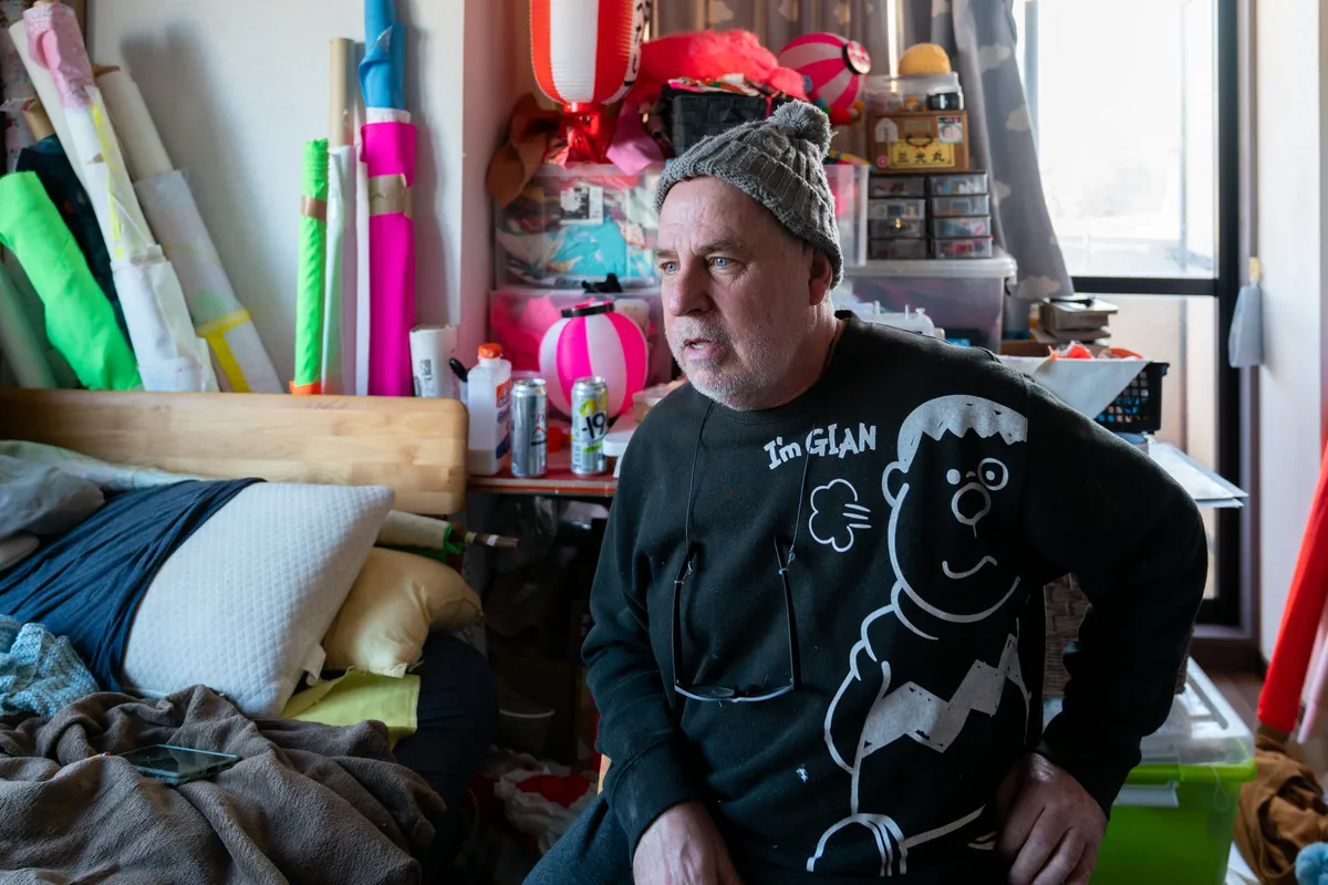 An environmental portrait of artist Dave Sayre in his cluttered workspace, surrounded by vibrant fabrics, Japanese lanterns, and various art supplies. The photograph captures a sense of creative immersion and the cross-cultural influences inherent in his life and work in Japan.