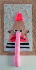 This mixed-media textile work features a stylized, slightly morose character with a long, stuffed pink nose and a cigarette, constructed from felt and faux fur. The central figure is set against a kitschy background of rainbow and unicorn patterns, creating a playful yet subversive contrast. The piece blends elements of soft sculpture and pop art to explore contemporary themes through a craft-oriented lens.