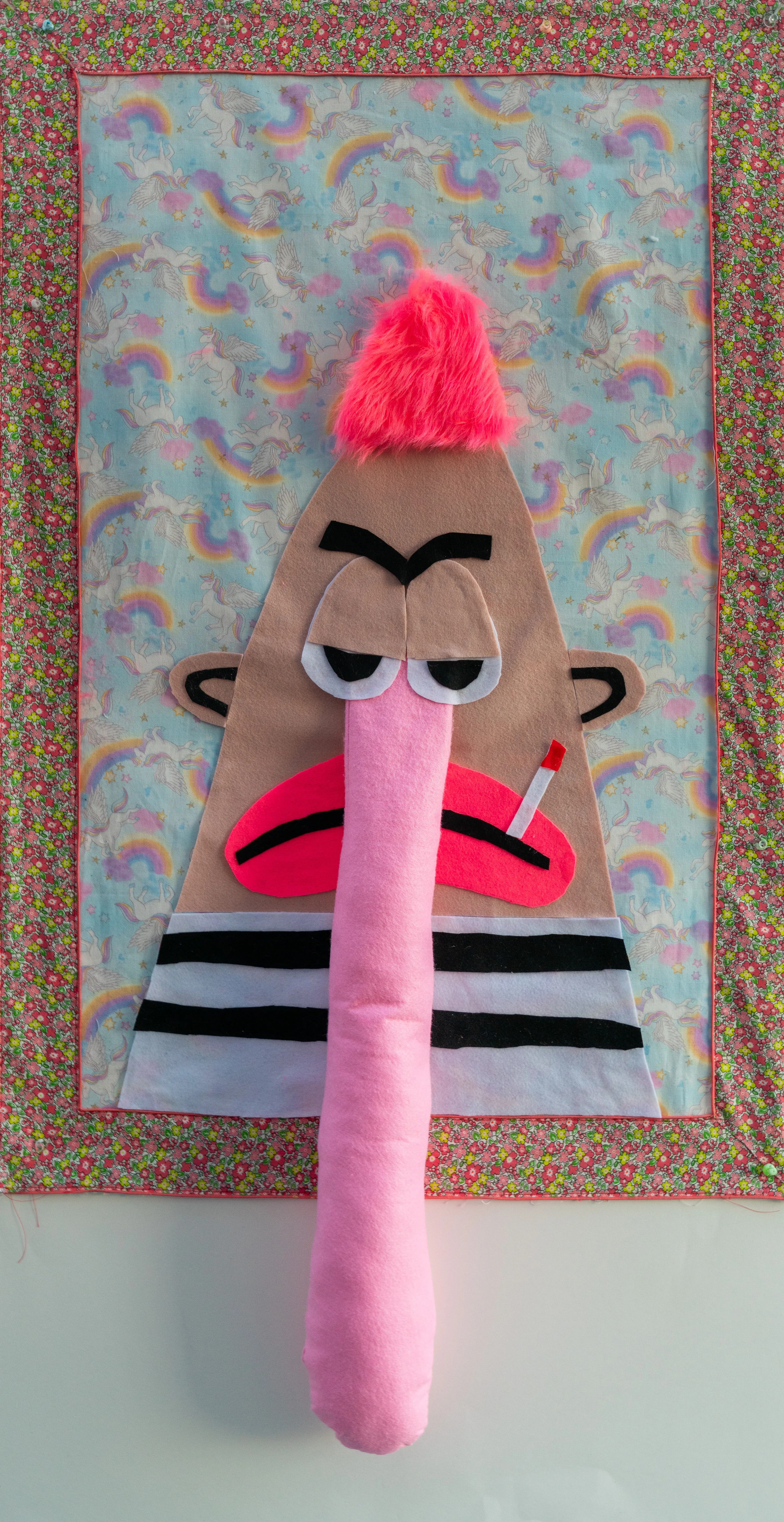 This tactile textile collage features a weary character with a plush, elongated nose and neon pink hair set against a whimsical background of rainbows and unicorns. The piece masterfully blends kitsch aesthetics with a sense of modern disillusionment through its use of soft fabrics and irreverent imagery. Sayre explores cross-cultural pop sensibilities using unconventional materials to create a bold, wall-mounted presence.